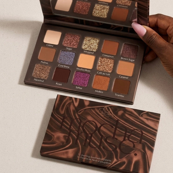 A makeup palette with multiple eyeshadow pans in warm neutrals, browns, and shimmery finishes, shown open with a hand holding one side. The closed palette is placed below it on a light beige surface.